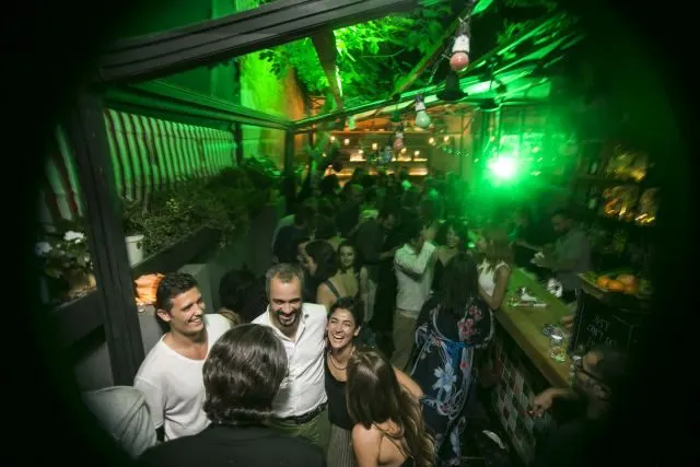Kapalı bir mekânda kalabalık insan topluluğu. Güçlü yeşil neon ışıklandırma ortamı aydınlatıyor. Tavanda asılı dekoratif elementler ve yeşil spot ışıklar. Ön planda genç insanlar birbirlerine yakın, eğlenirken. Arka planda daha fazla misafir ve bar tezgahı görülüyor. — KiKi, Sıraselviler 42, Cihangir, İstanbul