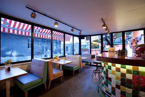 Geniş camlı modern bar. Pembe-beyaz çizgili toldolar pencerelerini dekore ediyor. Açık yeşil ve kahverengi deri banquette oturma alanları. Sıcak spot ışıklar tavan rayında. Sağda rengarenk mozaik karo bar tezgahı. Arka planda led ışıklı dekoratif elemanlar. — KiKi, Sıraselviler 42, Cihangir, İstanbul