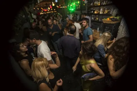 Dar, loş bir mekanın içinde yoğun kalabalık. Yeşil neon ışık ve sıcak sarı aydınlatma. İnsanlar birbirine yakın dans ediyor, müzik dinliyor. Arka planda bar tezgahı ve şişeler görünüyor. — KiKi, Sıraselviler 42, Cihangir, İstanbul