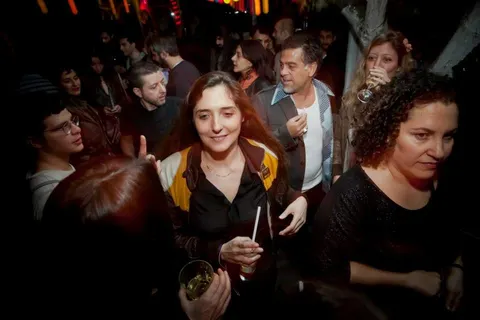 Kalabalık bir underground barda, sarı-siyah ceket giymiş kırmızı saçlı kadın ön planda içki tutarak gülümsüyor. Çevresinde müzik yapan ve eğlenen insanlar, sıcak turuncu-kırmızı aydınlatma altında dans ediyorlar. — KiKi, Sıraselviler 42, Cihangir, İstanbul