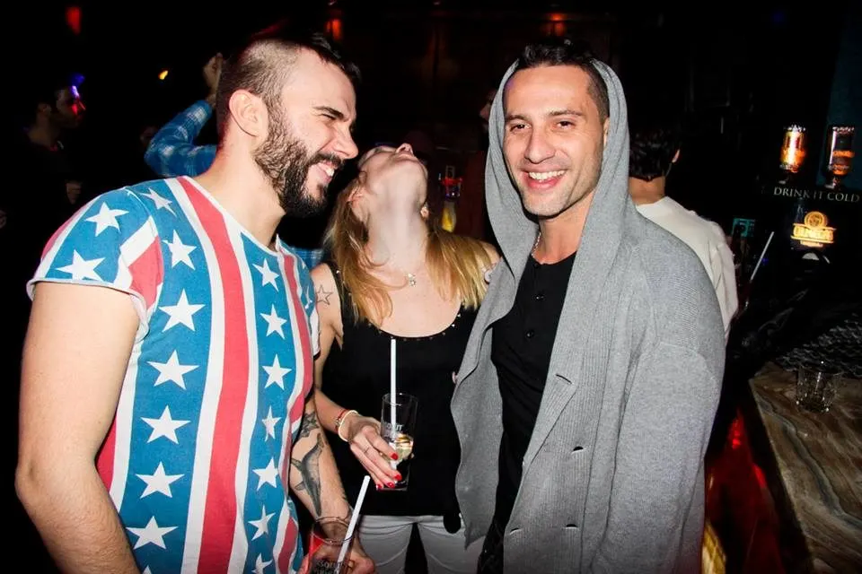 Gece barında üç kişi yan yana durarak sohbet ediyor. Soldaki erkek mavi-kırmızı yıldızlı tişört giyiyor, sağdaki erkek gri hoodie ile siyah tişört kombininde. Ortadaki kadın siyah giysi ve içki tutmuş. Arka planda kızıl-turuncu aydınlatma ve bar ortamı görülüyor. — KiKi, Sıraselviler 42, Cihangir, İstanbul