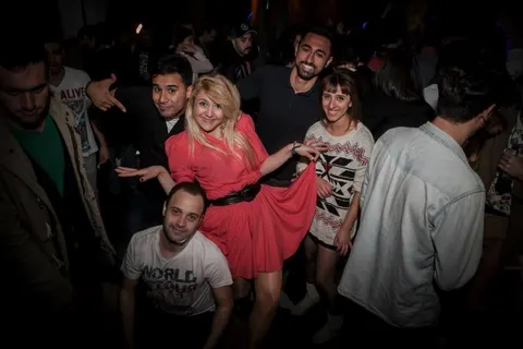 Karanlık mekânda canlı müzik eşliğinde dans eden ve sohbet eden grup. Pembe elbiseli kadın merkez konumdadır. Çeşitli kıyafetler giymiş erkekler ve kadınlar etrafında. Loş ışıklandırma, canlı bar atmosferi. — KiKi, Sıraselviler 42, Cihangir, İstanbul