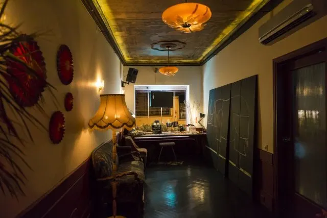 Dar koridorlu bar, sarı tonlu ışıklandırma, tavan aydınlatması, duvarlarda kırmızı dekoratif çiçekler, sağda koyu kapılar, solda sandalye ve dekorasyon, arka planda bar tezgahı görülüyor. — KiKi, Sıraselviler 42, Cihangir, İstanbul