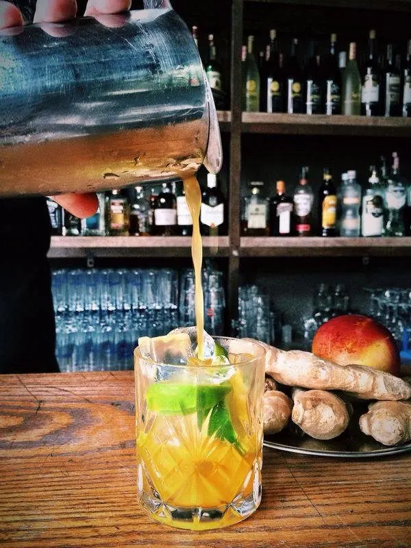 Barista bir bardağa amber renkli içecek döküyor. Ön planda buz dolu bardak, yeşil nane yaprağı, taze zencefil ve kırmızı elma görülüyor. Arka planda raflarda çeşitli şişeler dizili. — KiKi, Sıraselviler 42, Cihangir, İstanbul