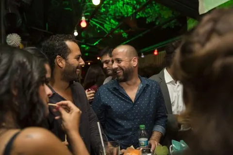Gece mekanında arkadaşlar sohbet ediyor. Yeşil ve kırmızı neon ışıklar tavan ve arka planda parlıyor. Ön planda mavi gömlekli adam, etrafında siyah kıyafetli kadınlar ve erkekler duruyor. Ellerinde bira şişeleri ve bardaklar var. Eğlenceli, samimi bir ortam. — KiKi, Sıraselviler 42, Cihangir, İstanbul
