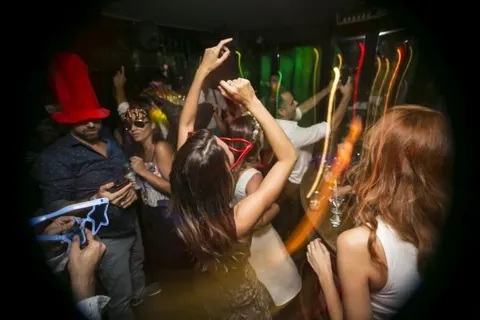 Karanlık mekanda renkli ışıklar arasında insanlar dans ediyor. Kırmızı şapkalı kişi, maskeliler ve ellerini havaya kaldıran dansçılar görülüyor. Arka planda gitar ve müzik enstrümanları ışıklandırılmış. — KiKi, Sıraselviler 42, Cihangir, İstanbul