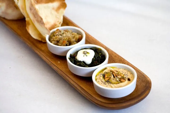 Ahşap uzun tahtaya dizilmiş üç beyaz kasede farklı mezze çeşitleri. Arka planda tostolmuş ekmek dilimleri. Kaseların içinde tahıllı salata, yeşil sebzeli beyaz peynir ve humus görülüyor. — KiKi, Sıraselviler 42, Cihangir, İstanbul
