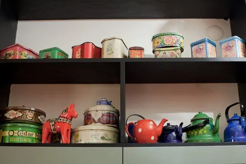 Siyah raflar üzerinde renkli metal kutular, seramik çaydanlıklar ve dekoratif nesneler. Kırmızı puantiyeli demitasse, yeşil çaydanlık, vintage pasta kutuları, çiçek desenli kaplar ve mavi emaye ketil. — KiKi, Sıraselviler 42, Cihangir, İstanbul