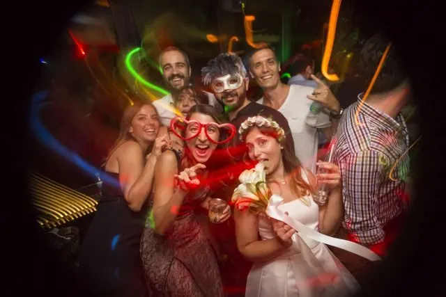 Karanlık barın ortasında sekiz kişi renkli neon ışıkları altında poz veriyor. Kırmızı ve beyaz kostümlü kadınlar maske takıyor, erkekler göz maskesi giyiyor. Yeşil, turuncu, mavi neon çizgiler arka planda hareket ediyor. — KiKi, Sıraselviler 42, Cihangir, İstanbul