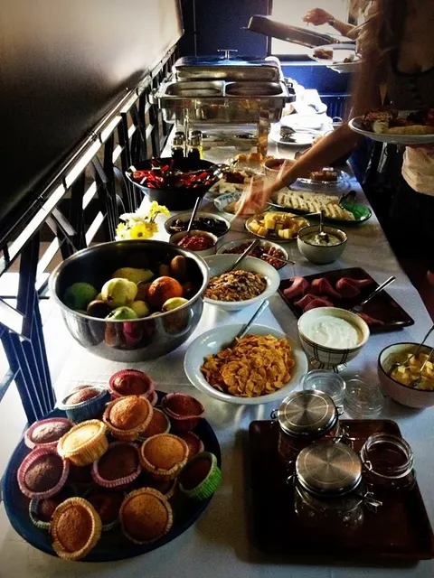 Uzun beyaz masada çok sayıda yemek dizili. Ön planda pasta ve tatlılar, metal kase içinde meyveler, beyaz tabakta salata ve sos, kırmızı et parçaları, kavanozlar, arka planda metal ısıtıcı kaplar ve insan elleri görülüyor. — KiKi, Sıraselviler 42, Cihangir, İstanbul