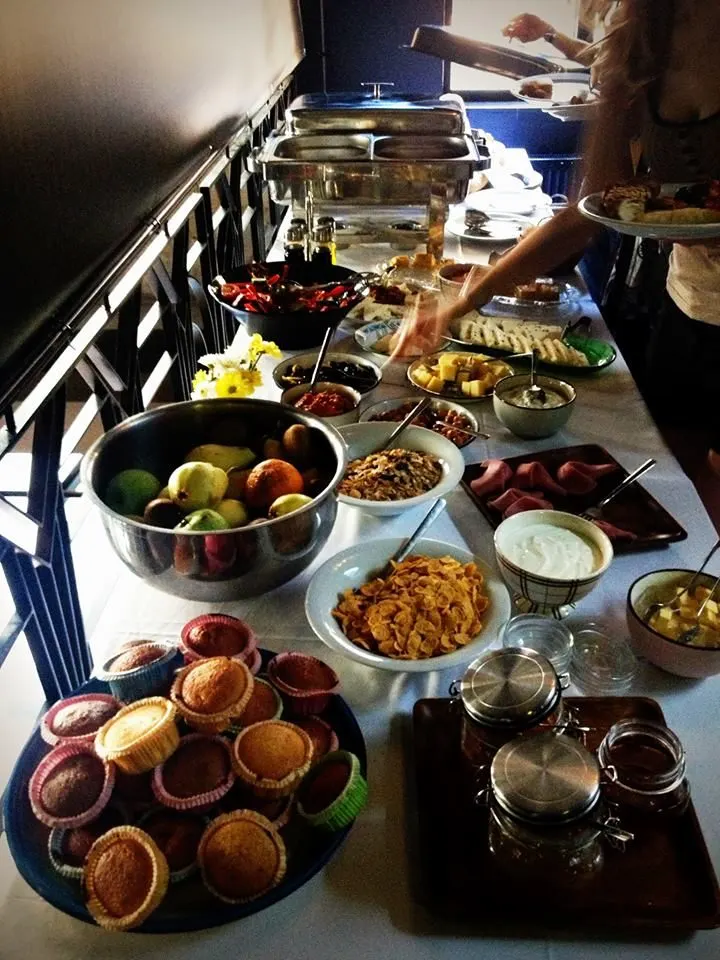 Uzun beyaz masada çok sayıda yemek dizili. Ön planda pasta ve tatlılar, metal kase içinde meyveler, beyaz tabakta salata ve sos, kırmızı et parçaları, kavanozlar, arka planda metal ısıtıcı kaplar ve insan elleri görülüyor. — KiKi, Sıraselviler 42, Cihangir, İstanbul
