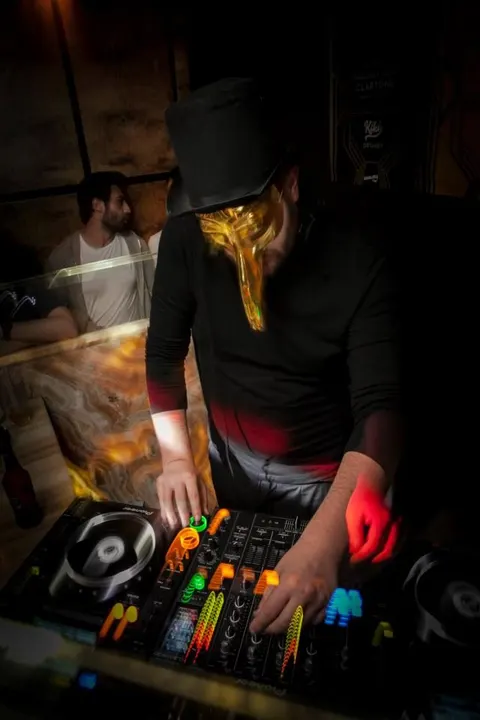 Siyah şapkalı DJ, neon sarı ve kırmızı ışıklar altında kontrolü elinde tutuyor. Ön planda renkli fader ve butonlar parlıyor. Arkada beyaz tişörtlü kişi gözlemliyor. Karanlık mekân, müzik enerjisi hissediliyor. — KiKi, Sıraselviler 42, Cihangir, İstanbul