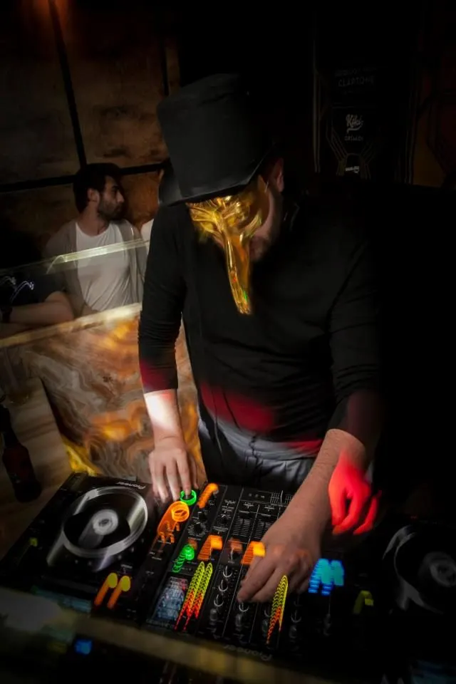 Siyah şapkalı DJ, neon sarı ve kırmızı ışıklar altında kontrolü elinde tutuyor. Ön planda renkli fader ve butonlar parlıyor. Arkada beyaz tişörtlü kişi gözlemliyor. Karanlık mekân, müzik enerjisi hissediliyor. — KiKi, Sıraselviler 42, Cihangir, İstanbul