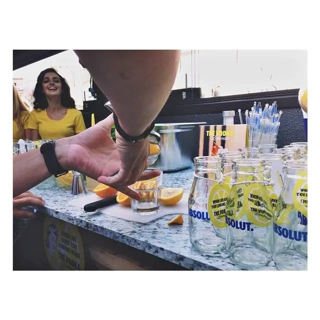 Mermer tezgahta limonlu içkiler ve bardaklar. Bir kişi limon dilimi ile bardağı tutuyor. Arka planda sarı tişörtlü kadın gülümsüyor. Parlak gündüz aydınlatması. Cam şişelerde sarı etiketli Absolut likörü. — KiKi, Sıraselviler 42, Cihangir, İstanbul