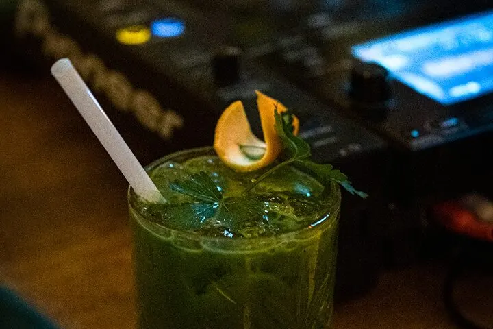 Bulanık arka plan önünde, buz dolu yeşil renkli kokteyl bardağı. Beyaz pipet ve taze mint yapraklığı içinde. Üstünde sarı limon dilimi dekorasyonu. Koyu ortamda ışık parıltıları görülüyor. — KiKi, Sıraselviler 42, Cihangir, İstanbul