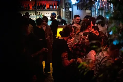 Sıcak turuncu ve sarı ışıklandırmanın aydınlattığı bar içinde yoğun kalabalık. Ön planda insanlar sıkı sıkıya yan yana duruyor. Arka planda bardaki şişeler ve ışıklar görülüyor. Rahat, canlı bir gece hayatı ortamı. — KiKi, Sıraselviler 42, Cihangir, İstanbul
