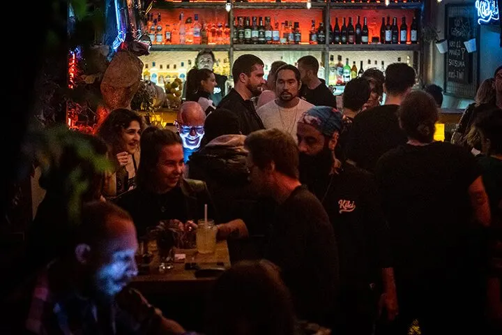 Sıcak turuncu ve mavi neon ışıkların aydınlattığı bar ortamında yoğun kalabalık. Arka planda şişe rafları ve aydınlatılmış duvar. İnsanlar sıkı sıkıya bir araya toplanmış, bazıları konuşuyor, bazıları içki tutuyor. — KiKi, Sıraselviler 42, Cihangir, İstanbul