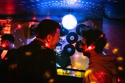 Karanlık mekanın içinde insanlar durmuş, mavi ve kırmızı neon ışıklar çevreyi aydınlatıyor. Üst tarafta parlak beyaz ve mavi led paneller var. Arka planda dj kulaklıkları olan bir portre görülüyor. Sıcak sarı ve turuncu ışıklar da mekanı renklendiriyor. — KiKi, Sıraselviler 42, Cihangir, İstanbul