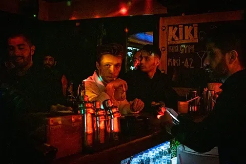 Karanlık bar ortamında, kırmızı-turuncu aydınlatma altında beş erkek müşteri ve barista görülüyor. Masada şişeler, bardaklar ve içecekler dizili. Duvar üzerinde mavi ışıklı işaretler vardır. — KiKi, Sıraselviler 42, Cihangir, İstanbul