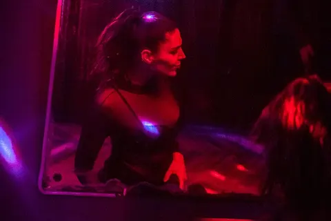 Kırmızı ve mor neon ışıklandırmayla aydınlatılan karanlık bir barın içinde, DJ booth'un arkasında bir kadın DJ görülüyor. Sıcak kırmızı ve soğuk mor ışıklar mekanın duvarlarına yansıyor. — KiKi, Sıraselviler 42, Cihangir, İstanbul