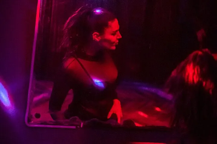 Kırmızı ve mor neon ışıklandırmayla aydınlatılan karanlık bir barın içinde, DJ booth'un arkasında bir kadın DJ görülüyor. Sıcak kırmızı ve soğuk mor ışıklar mekanın duvarlarına yansıyor. — KiKi, Sıraselviler 42, Cihangir, İstanbul