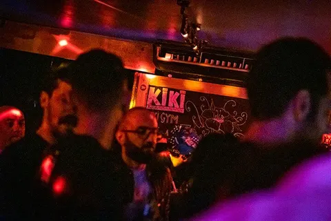 Kırmızı ve mor neon ışıklarıyla aydınlanan dar ve kalabalık bir mekan. Ön planda hareket halindeki insanların siluetleri, arka planda sarı neon tabelalar ve duvar dekorasyonları görülüyor. Canlı müzik ortamı, sıcak ve mat aydınlatma atmosferi. — KiKi, Sıraselviler 42, Cihangir, İstanbul