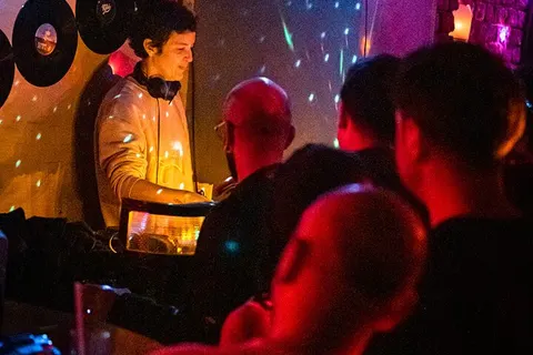 Siyah ışıklı underground bar ortamında, DJ booth'ta kulaklıkla duran DJ figure görülüyor. Önünde vinyl plaklar ve ekipman var. Kırmızı, sarı ve mavi renkli ortam ışıklandırması. Ön planda dans eden müzik severler kalabalığı. — KiKi, Sıraselviler 42, Cihangir, İstanbul