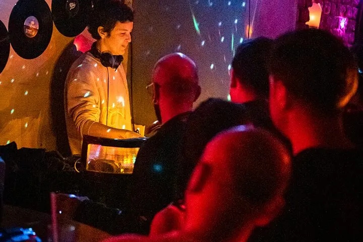 Siyah ışıklı underground bar ortamında, DJ booth'ta kulaklıkla duran DJ figure görülüyor. Önünde vinyl plaklar ve ekipman var. Kırmızı, sarı ve mavi renkli ortam ışıklandırması. Ön planda dans eden müzik severler kalabalığı. — KiKi, Sıraselviler 42, Cihangir, İstanbul