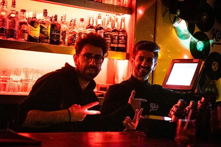 İki erkek barista karanlık barda kırmızı ışıkla aydınlanan tezgahın arkasında duruyor. Arkalarında cam raflarda likör şişeleri, yeşil neon trafik ışığı görülüyor. Tezgahın üstünde tablet bilgisayar var. — KiKi, Sıraselviler 42, Cihangir, İstanbul