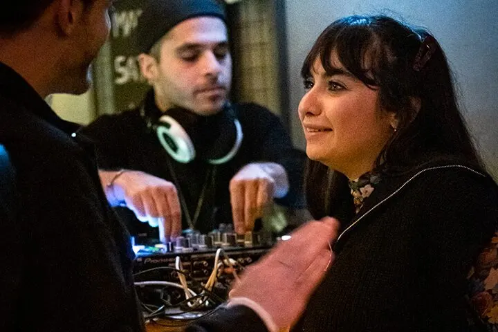 Karanlık mekânda, aydınlık turntable başında insanlar sohbet ediyor. Ön planda bir kişinin eli görülüyor, ortada başlık takılı DJ, sağda uzun siyah saçlı kadın gülümsüyor. Mavi ışık sarı dans pisti ekipmanını vurguluyor. — KiKi, Sıraselviler 42, Cihangir, İstanbul