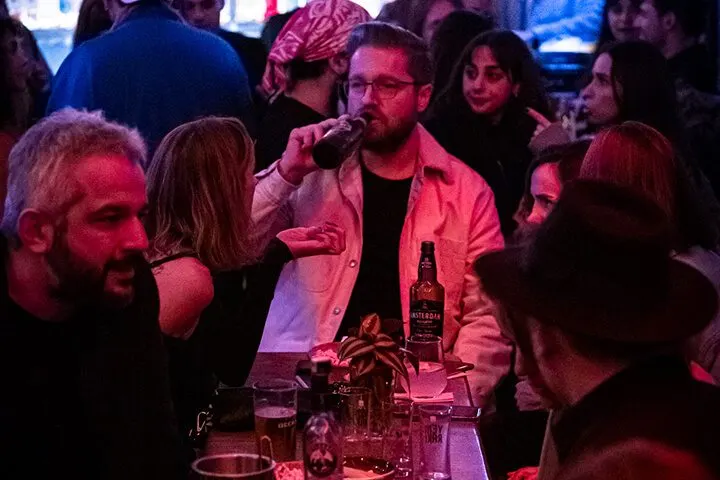 Pembe ve mor ışıklandırmalı bar ortamında, masada oturan erkek mikrofonla konuşuyor. Etrafında birkaç kişi vardır. Masada içki şişeleri ve bardaklar görülmektedir. Arkaplan kalabalık ve karanlık. — KiKi, Sıraselviler 42, Cihangir, İstanbul