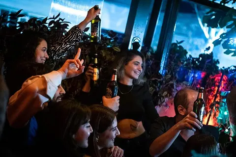 Neon ışıklarıyla aydınlanan bar ortamında, bir grup genç arkadaş bira şişeleriyle havaya kaldırarak kutlama yapıyor. Arka planda renkli LED ışıkları ve yeşil bitkiler görülüyor. — KiKi, Sıraselviler 42, Cihangir, İstanbul