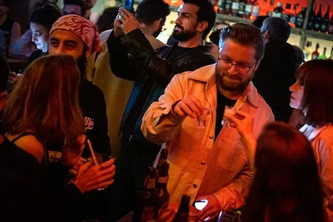 Sıcak turuncu ve amber ışıklar altında, gözlüklü orta yaşlı bir adam ön planda dans ederken, arkasında ve yanında çeşitli yaşlardaki insanlar müzik eşliğinde hareket ediyor. Bar atmosferi, kalabalık ve canlı. — KiKi, Sıraselviler 42, Cihangir, İstanbul