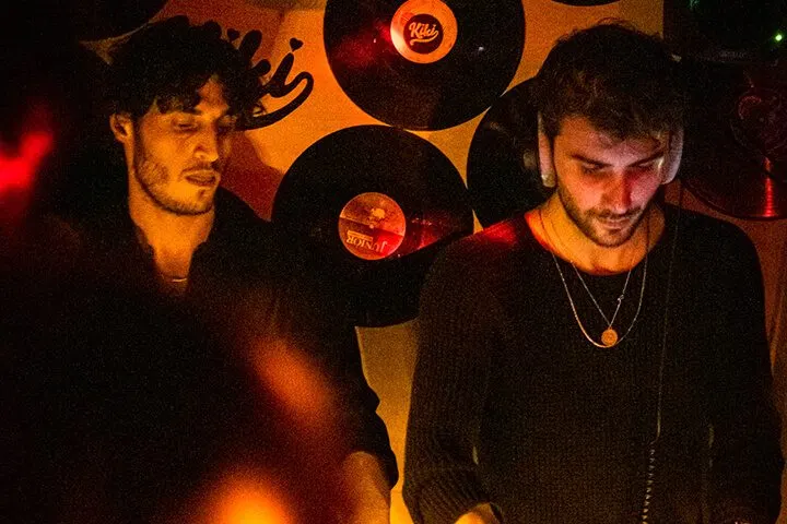 Siyah giyimli iki erkek DJ, turuncu ışıklandırma altında müzik setine konsantre. Arkalarında siyah vinilPlaklar görülüyor. Kırmızı ışık saçan lambalar ve karanlık ortam gece hayatı atmosferini oluşturuyor. — KiKi, Sıraselviler 42, Cihangir, İstanbul