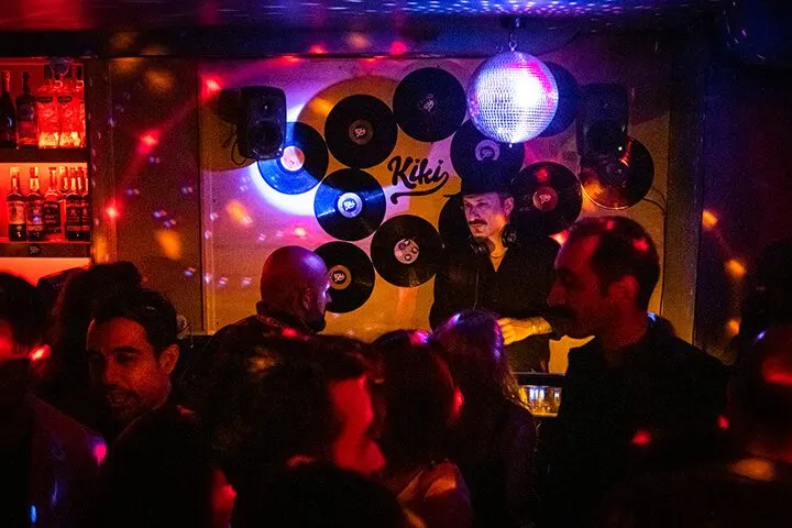 Karanlık bar ortamında mor, mavi ve kırmızı neon ışıklar yanıyor. Duvarı süsleyen siyah vinilplaklar arasında şapkalı DJ figürü görülüyor. Ön planda kalabalık insanlar dans ediyor, arka planda bar tezgahı aydınlık görünüyor. — KiKi, Sıraselviler 42, Cihangir, İstanbul