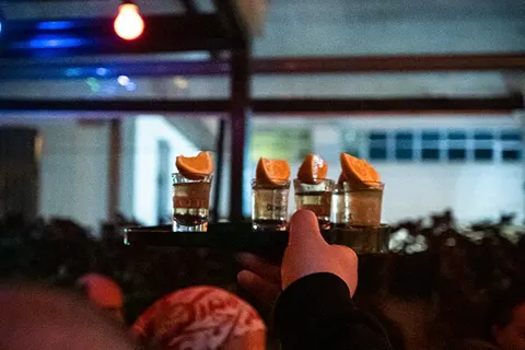 Koyu bar ortamında, aydınlık bardaklar önünde kalabalık içinde birinin eli uzanıyor. Bardaklarda portakal garnişleri var. Arka planda mavi ve kırmızı ışıklar, büyük pencereler görülüyor. — KiKi, Sıraselviler 42, Cihangir, İstanbul