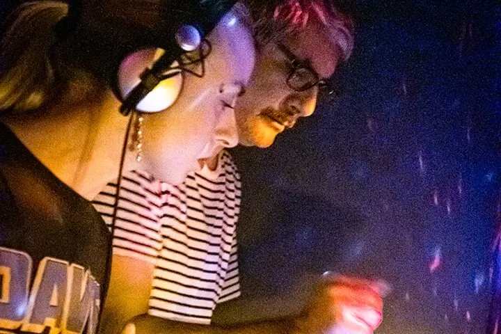 Gözlüklü DJ'nin yanından görünüş. Başında kulaklıklar, çizgili beyaz-siyah tişört giymiş. Mor ve sarı ışıklar arasında DJ setup'ında çalışıyor. Arka planda koyu, atmosferik gece mekânı ortamı. — KiKi, Sıraselviler 42, Cihangir, İstanbul