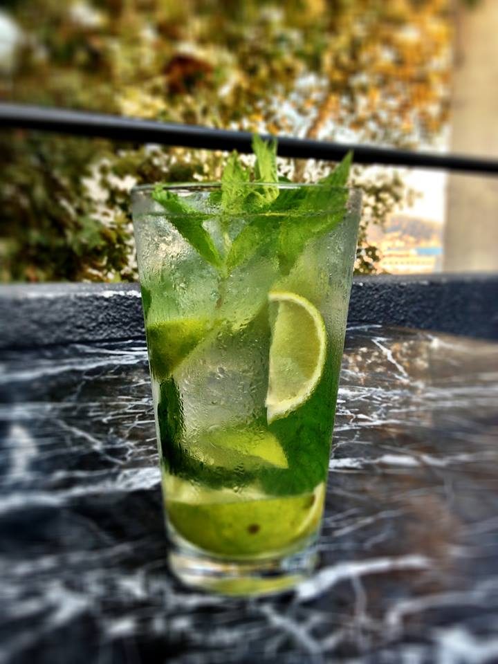 Buz dolu bardakta yeşil mojito kokteyli, taze nane yaprakları ve limon dilimleriyle garnili. Işıl ışıl buharlaşmış camda, bulanık bahçe ortamında güneş ışığı altında. — KiKi, Sıraselviler 42, Cihangir, İstanbul
