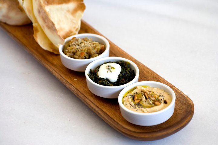 Ahşap uzun tahtaya dizilmiş üç beyaz kasede farklı mezze çeşitleri. Arka planda tostolmuş ekmek dilimleri. Kaseların içinde tahıllı salata, yeşil sebzeli beyaz peynir ve humus görülüyor. — KiKi, Sıraselviler 42, Cihangir, İstanbul