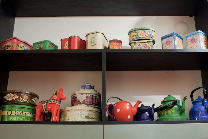 Siyah raflar üzerinde renkli metal kutular, seramik çaydanlıklar ve dekoratif nesneler. Kırmızı puantiyeli demitasse, yeşil çaydanlık, vintage pasta kutuları, çiçek desenli kaplar ve mavi emaye ketil. — KiKi, Sıraselviler 42, Cihangir, İstanbul