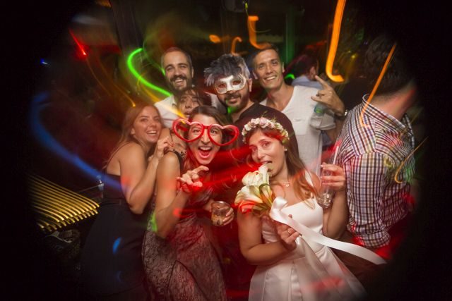 Karanlık barın ortasında sekiz kişi renkli neon ışıkları altında poz veriyor. Kırmızı ve beyaz kostümlü kadınlar maske takıyor, erkekler göz maskesi giyiyor. Yeşil, turuncu, mavi neon çizgiler arka planda hareket ediyor. — KiKi, Sıraselviler 42, Cihangir, İstanbul